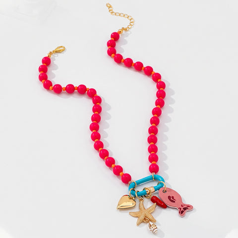 A beaded necklace with bright pink round beads, gold-tone chain and connectors, and three dangling charms: a turquoise heart, a gold starfish, and a pink fish, displayed on a clean white background.