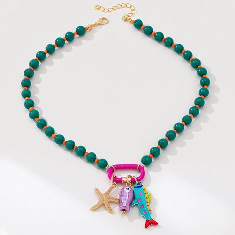 A beaded necklace with round green beads separated by small orange spacer beads, a gold-tone chain extender and clasp, and a cluster of colorful charms including a pink carabiner, a starfish, a purple fish, and a blue-green fish with a red tail, shown on a white background.