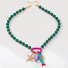 A beaded necklace with round green beads separated by small orange spacer beads, a gold-tone chain extender and clasp, and a cluster of colorful charms including a pink carabiner, a starfish, a purple fish, and a blue-green fish with a red tail, shown on a white background.
