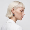 A close-up side profile of a model with short platinum blonde hair and freckles, wearing a black beaded drop earring with small white dots and a gold hoop, paired with a white collared shirt against a light gray background.