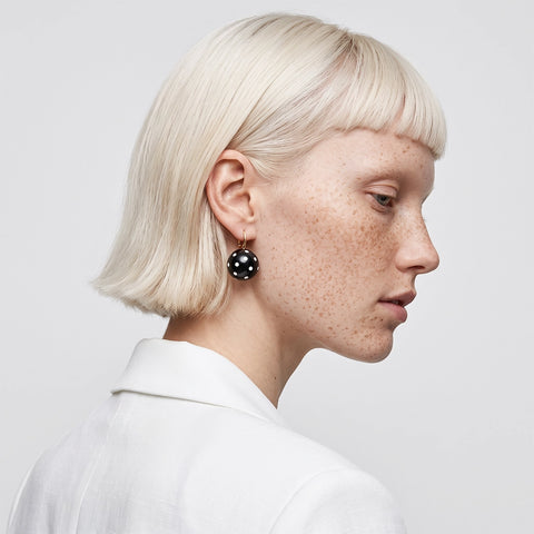 A close-up side profile of a model with short platinum blonde hair and freckles, wearing a black beaded drop earring with small white dots and a gold hoop, paired with a white collared shirt against a light gray background.