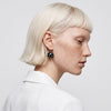 A close-up side profile of a model with short platinum blonde hair and freckles, wearing a black beaded drop earring with small white dots and a gold hoop, paired with a white collared shirt against a light gray background.