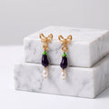 A pair of gold-tone dangle earrings shaped like eggplants, with dark purple bodies, small green tops, bow-shaped posts, and dangling white pearl beads, displayed on stacked white marble blocks against a light gray background.
