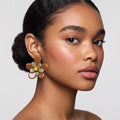 A close-up portrait of a model wearing a large flower-shaped earring. The earring features brown petals with olive green and pale yellow accents, set against a neutral light gray background.