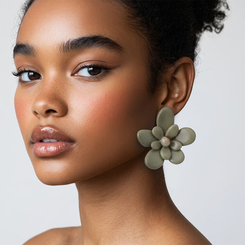 A close-up of a model wearing a large, sculptural olive green flower-shaped stud earring against a clean, light gray background.