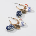 A pair of gold hoop earrings with dangling charms, including blue and white patterned ceramic beads, gold heart charms, and a small red and white bead, displayed on a light gray background.