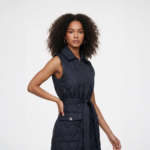 A woman modeling a sleeveless quilted navy puffer vest with a front zipper, collar, and matching tie belt, photographed against a clean light gray studio background.