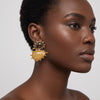 A close-up side profile of a model wearing a large, two-tier gold statement earring. The earring features a smaller dark brown teardrop stone on top and a larger, polished yellow-gold teardrop stone below, both set in a gold frame with small gold spikes.