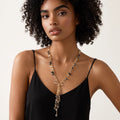 A woman with curly dark hair wearing a long gold-toned statement necklace with black and white shell beads, shown from the chest up against a plain light background.