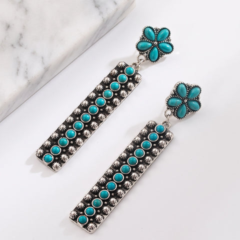 A pair of silver-toned dangle earrings with turquoise blue stone accents, displayed on a clean white surface with a marble block in the background.