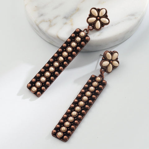 A pair of long rectangular drop earrings with a dark brown textured body, lined with cream-colored oval stones and small starburst accents, topped with a matching cream flower-shaped stud, displayed on a white marble slab against a white background.