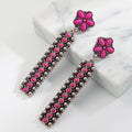 A pair of dangling statement earrings with bright pink flower-shaped studs and long rectangular drops covered in pink and silver-toned embellishments, displayed on a white marble block against a white background.