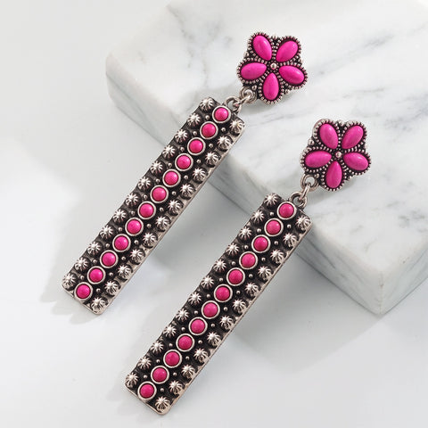 A pair of dangling statement earrings with bright pink flower-shaped studs and long rectangular drops covered in pink and silver-toned embellishments, displayed on a white marble block against a white background.