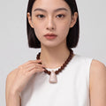 A woman wearing a dark brown beaded necklace with a light pink teardrop-shaped pendant, paired with a white sleeveless top against a light gray background.