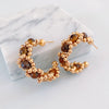A pair of gold hoop earrings with textured beaded details and brown tiger's eye stones, displayed on a white marble block against a light gray background.