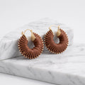 A pair of large gold hoop earrings with textured, pleated brown circular dangles, displayed on a white and gray marble block against a clean white background.